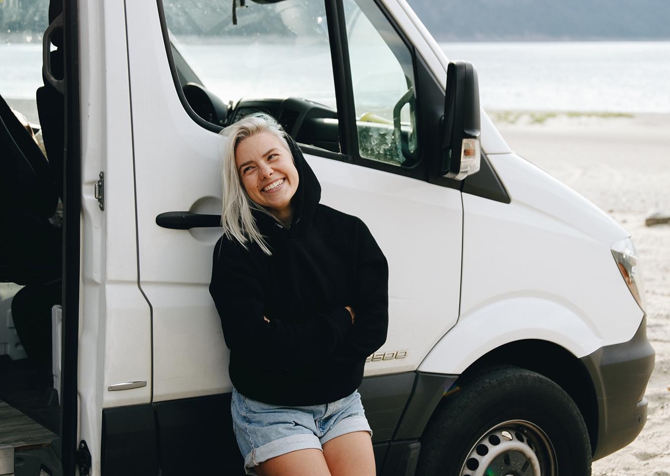 Glad kvinna i huvtröja och korta shorts lutar sig vi dörren på sin husbil