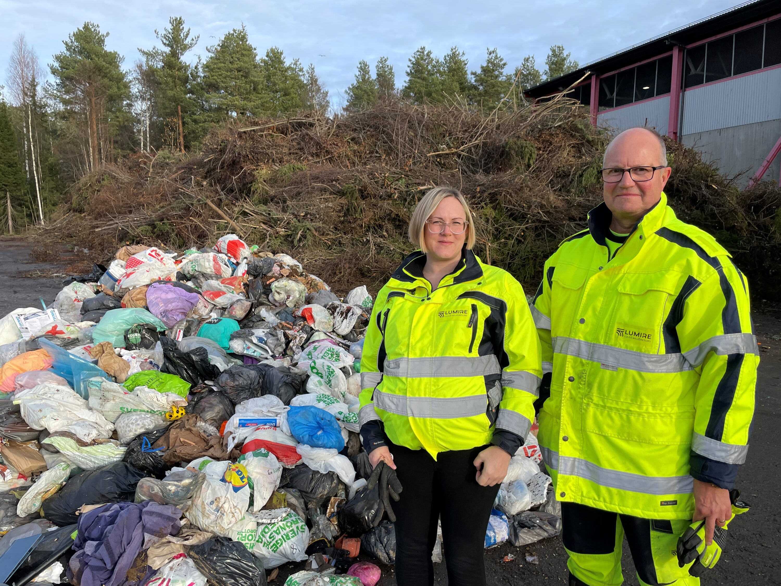 Två personer i gula arbetskläder står framför en hög av sopsäckar.