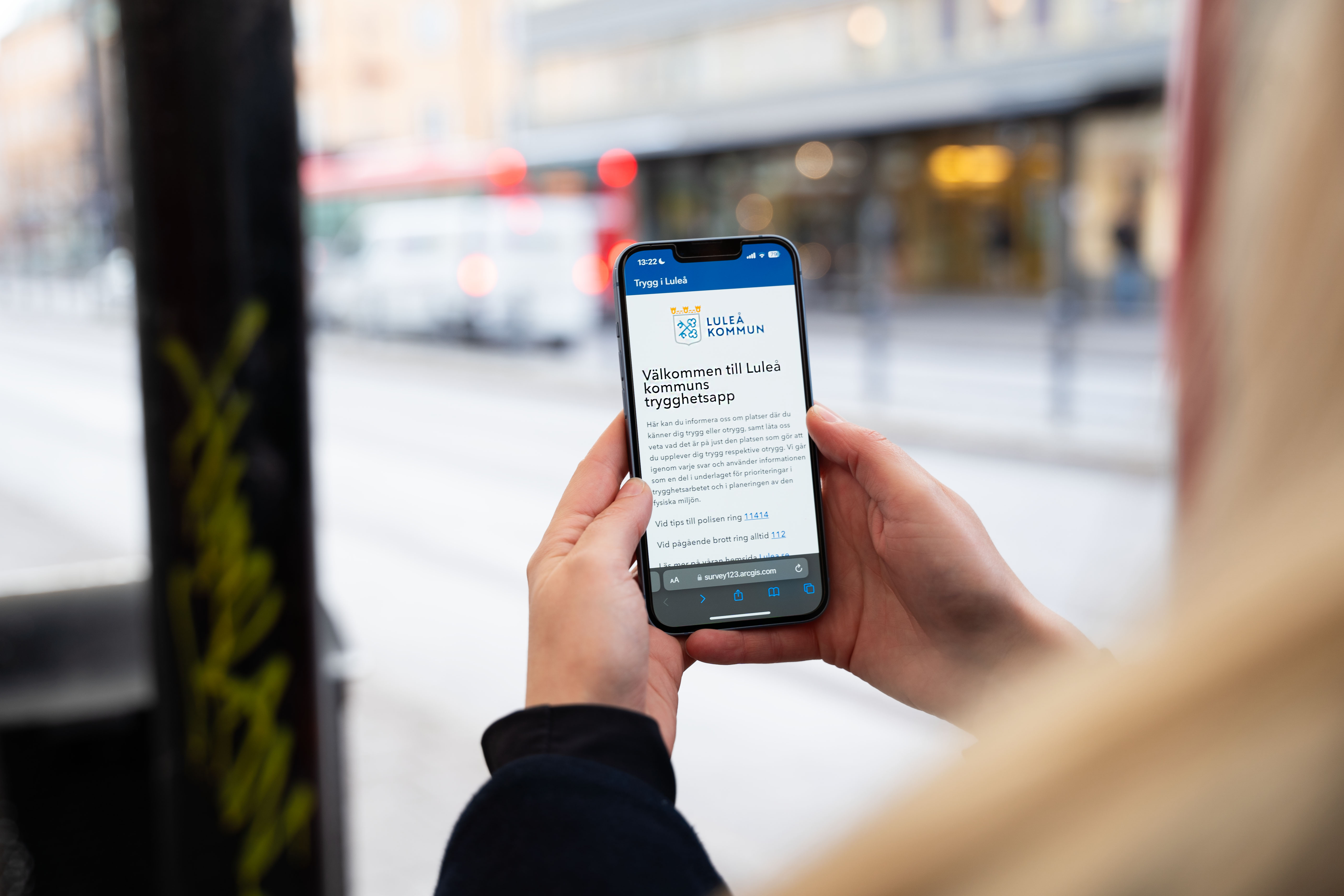 En person håller i en mobiltelefon. Personen befinner sig på Smedjegatans busstation. På mobilskärmen syns verktyget "Trygg i Luleå".