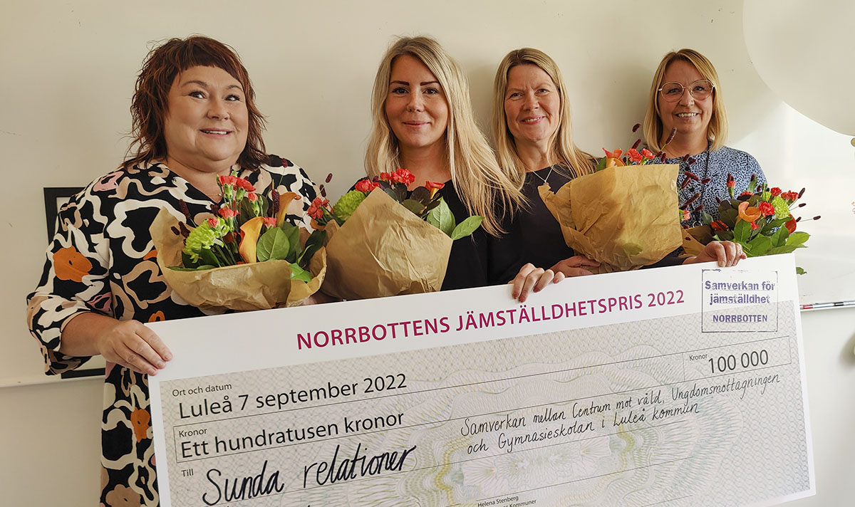 Fyra glada kvinnor håller i en stor check, och blommor.