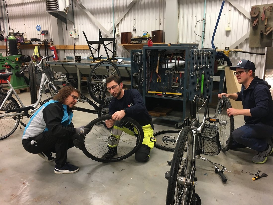Två personer sitter på huk och byter ett cykeldäck och i närheten tar en annan loss ett däck från en cykel. En massa verktyg i bakgrunden.