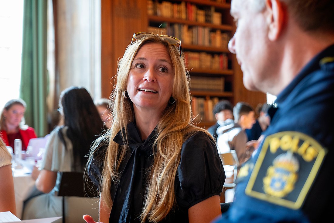 Åsa Koski sitter vid ett bord och pratar med en man i Polisuniform. 