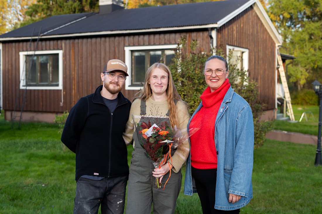 Tre personer står framför ett litet, brunt trähus.